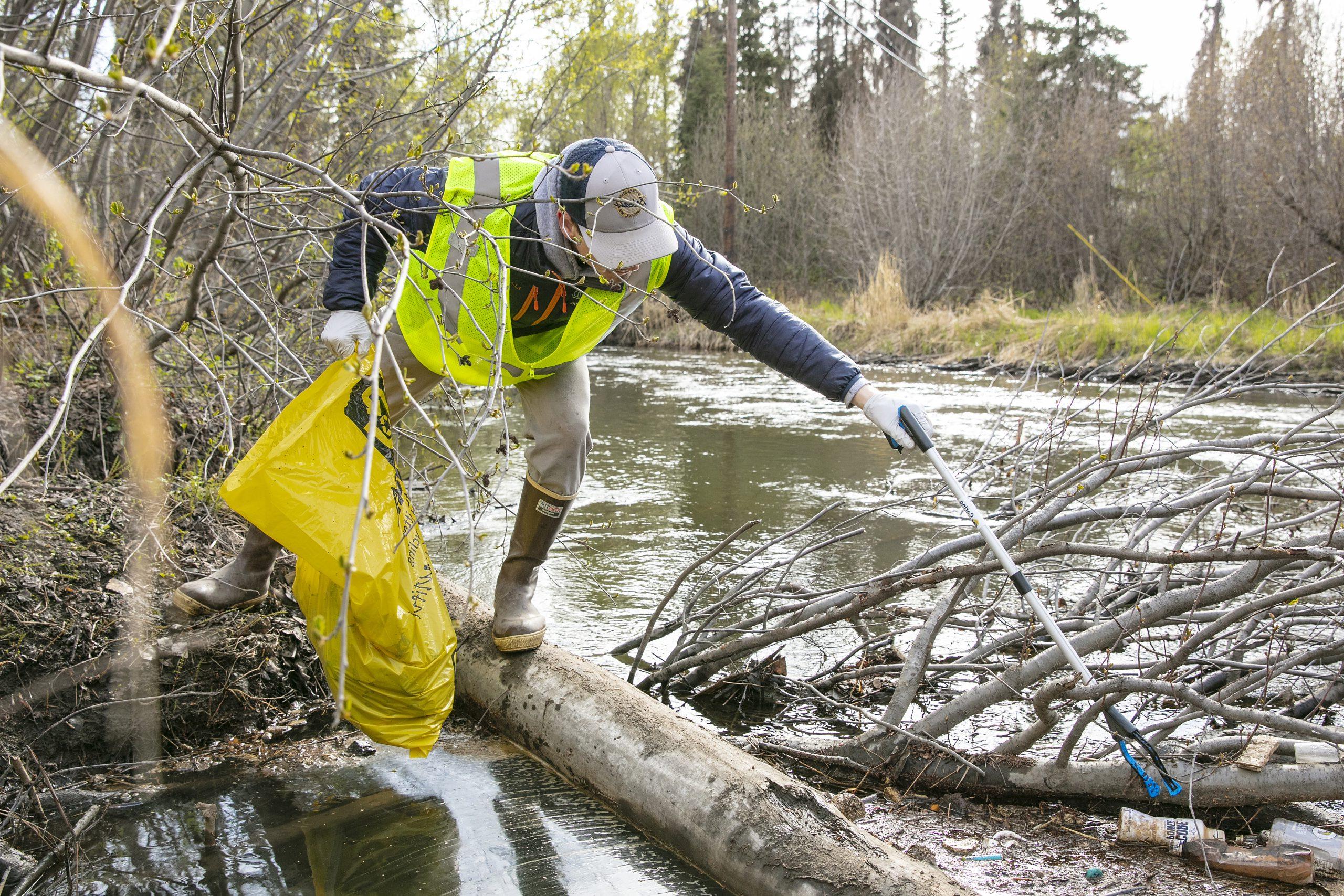 A volunteer wearing rubber boots and yellow safety vest holds a yellow trash bag while reaching with a grabber to pick up trash in Campbell Creek.